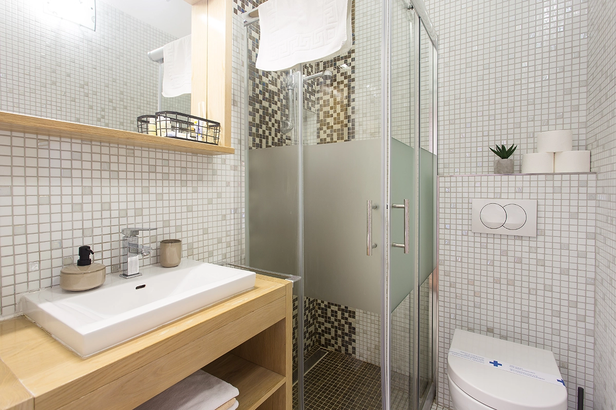 Modern bathroom connected to the room, with small mosaic tiles, a shower cabin, toilet, sink, and wooden cabinets.